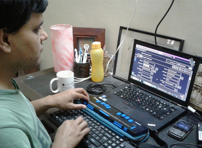 Pradip Sinha, a man who is deaf blind sits at a table with one hand on a keyboard and the other on a refreshable braille display. A laptop and cellphone are seen in front of him.
